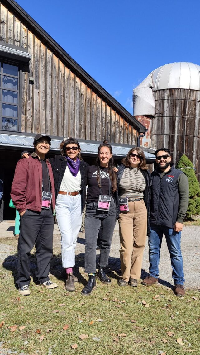 Photo of 5 photojournalists standing side by side in front of a brown shingled barn smiling.