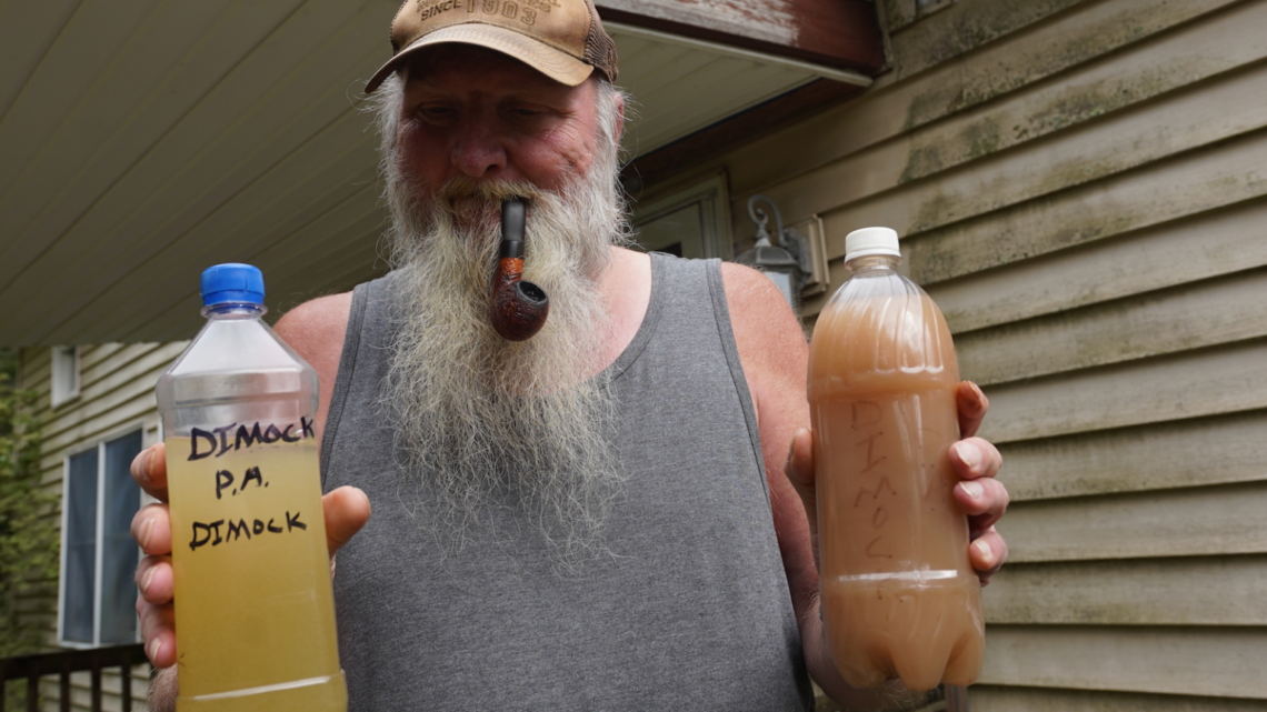 Ray Kemble stands with a tobacco pipe in his mouth holding two bottles of yellow and brown water from his home in two plastic water bottles.