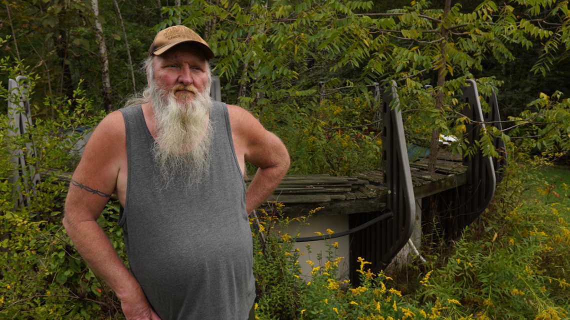 Ray Kemble, a white man with a long white beard, is wearing a gray tank top and a brown cap, and standing in front of overgrown foliage in front of his unusable swimming pool. 