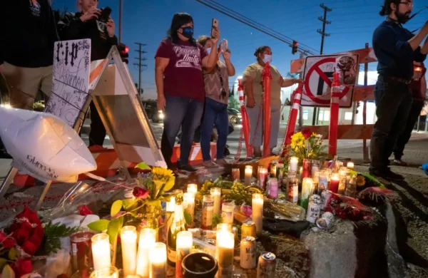 Mourners stand near a streetside memorial with candles and flowers."