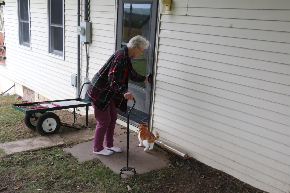 Mary Ellen McConnell stands outside with her cat. She is using a walker with one hand and opening the door to the house with the other. 