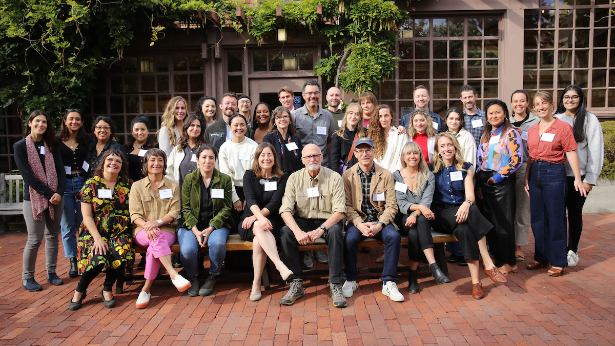 A large group of people stand and sit in front of a rustic building with glass-paned windows, giving it a home-like charm. Some are smiling while others look serious, dressed in a mix of casual and business attire with name tags pinned to their clothes. They gather on a red brick patio.
