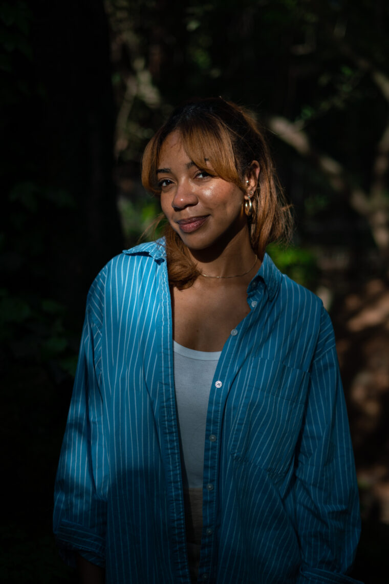 Photo of producer Myah Overstreet wearing a blue button down shirt in partial profile in front of a wooded background. 