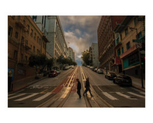 Two people cross a street as evening approaches in an urban environment. Steep incline flanked by tall buildings; rails embedded in the road hint at cable car transit. Cloudy sky above, street signs partly visible, cars parked along the sidewalk—captured for a student photography exhibit.
