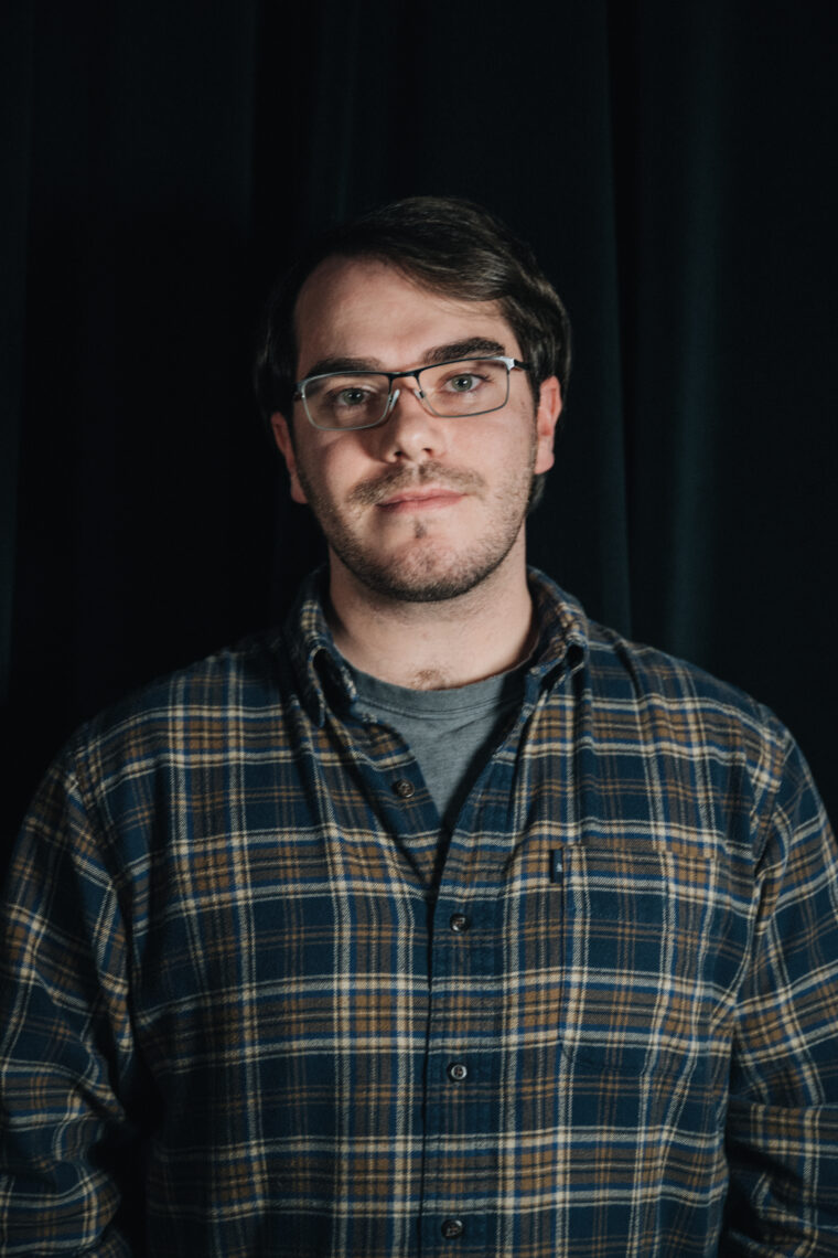 Samuel Tanner. A Berkeley journalism student looking into the camera. He has short brown hair, is wearing glasses, and a plaid shirt with a grey t-shirt underneath. 