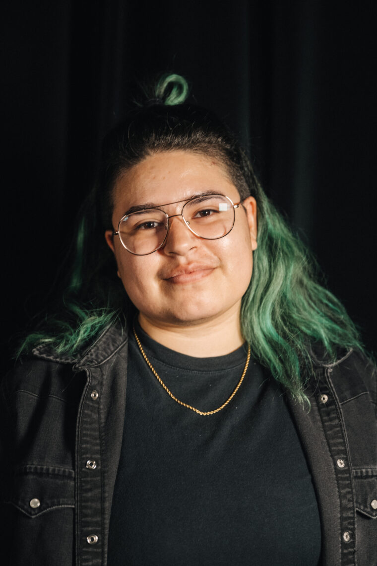 A woman with glasses is posed in front of the camera. Her hairstyle is in a half up half down look with green ends. She is wearing a plain black shirt with a black denim button-up shirt and a gold chain. The background is plain black. 