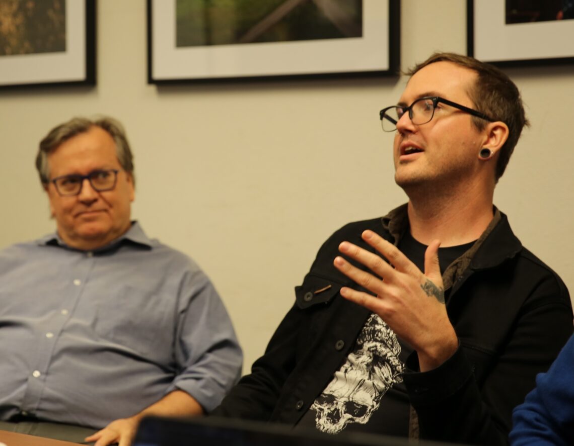 Two men are sitting and engaging in a discussion. The man on the right, wearing glasses and a black jacket, gestures with his hand while speaking. The man on the left, in a blue shirt, listens attentively. Framed pictures hang on the wall behind them.