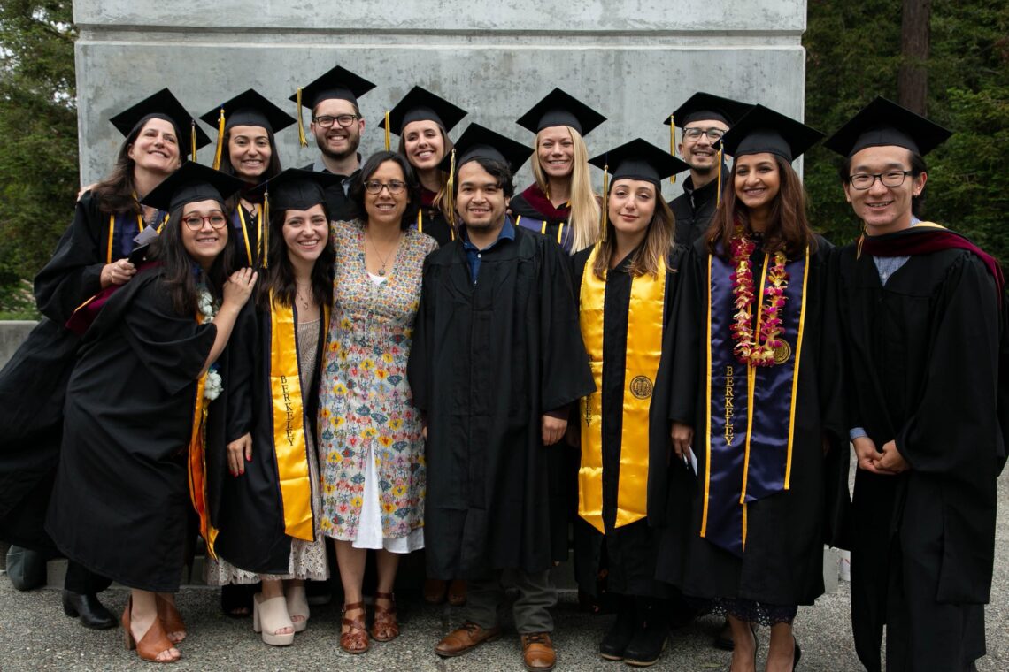 A group of people in graduation attire stands together in front of a concrete structure. Some graduates wear black gowns and caps with tassels and yellow stoles. They smile at the camera, flanked by two individuals in everyday clothing. Trees are visible in the background as filmmaker Carrie Lozano delivers the commencement keynote.