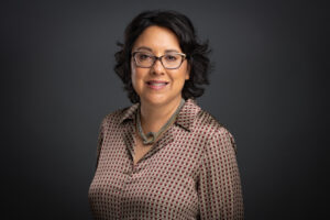 A person with short, dark hair and glasses smiles slightly, wearing a patterned blouse and a chunky necklace. The backdrop is a dark, plain color, setting the perfect stage for filmmaker Carrie Lozano as she prepares to deliver her commencement keynote.