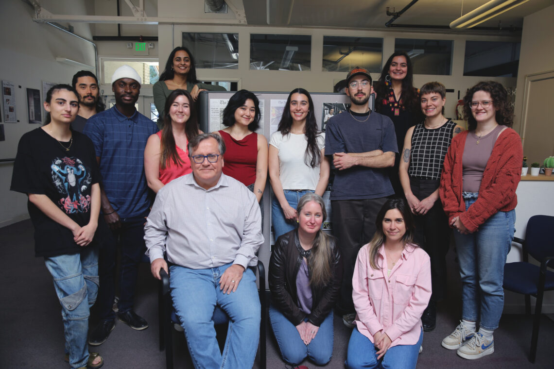 A group of 15 people posing for a photo in an office setting, some standing while others are seated. The group includes a mix of men and women of various ages, dressed casually. Dean Geeta Anand is among them. The background features office furnishings and wall decorations, perfect for the Quarterly Newsletter.