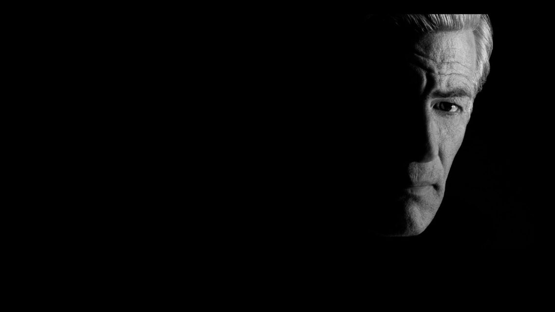 A grayscale image shows the right half of an older man's face emerging from a pitch-black background. His expression is somber and thoughtful, with deep shadows accentuating his features, including furrowed brows and faint wrinkles on his forehead—a powerful capture reminiscent of James Nachtwey's work.