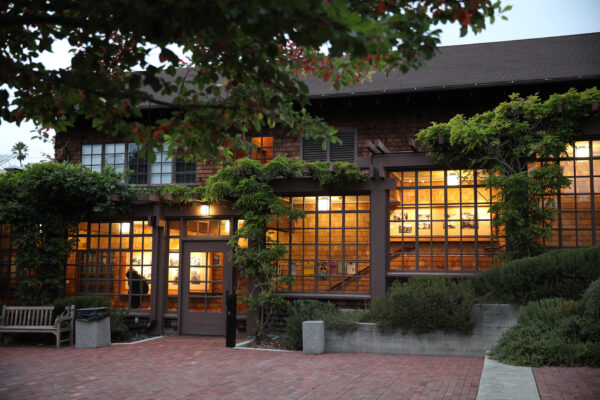 A cozy building with large illuminated windows at dusk. The exterior features brick and wooden elements with lush greenery climbing the facade. A brick walkway leads to the entrance, where advisors often meet on a wooden bench surrounded by small plants near the doorway.