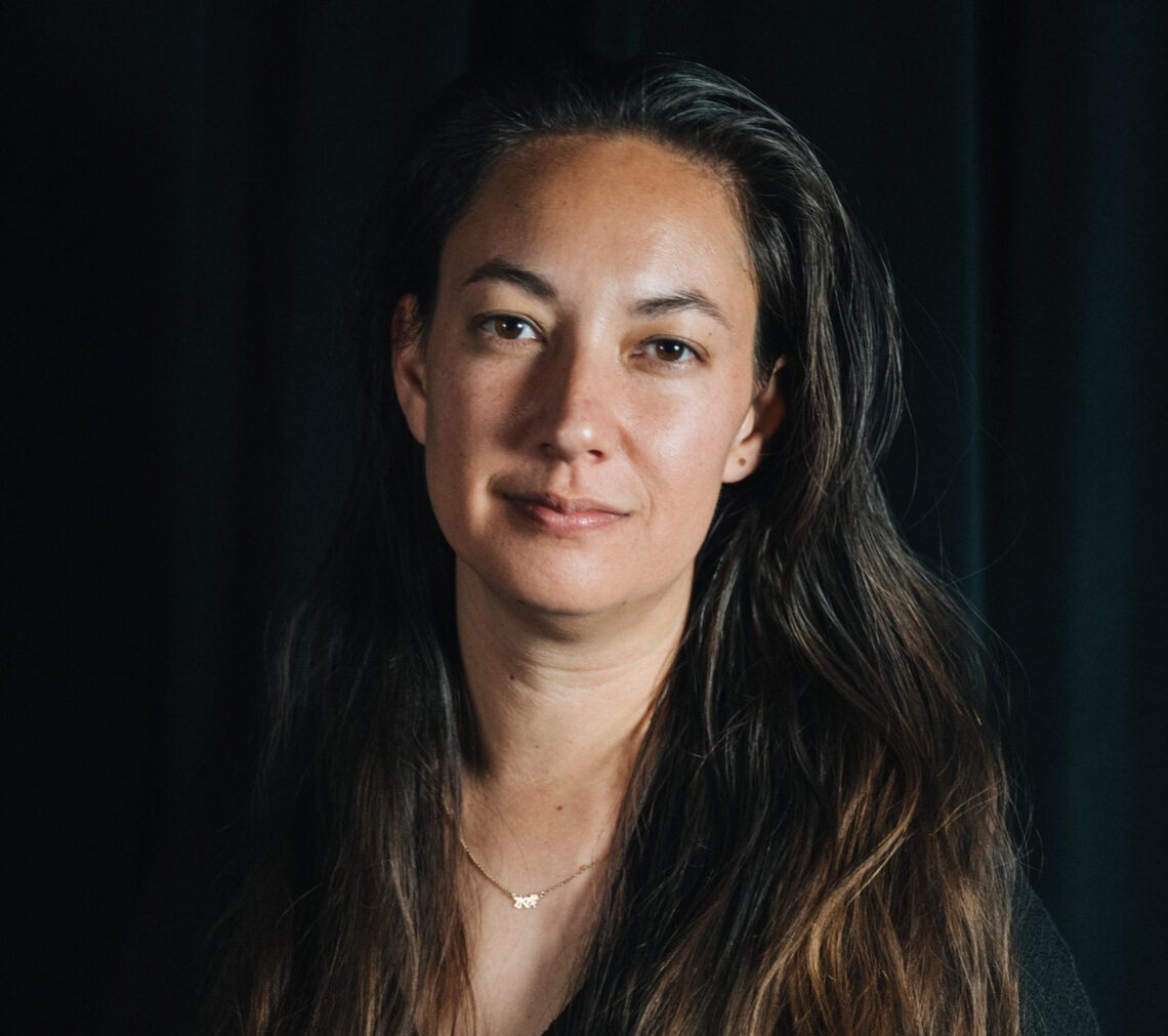 A woman with long, wavy, dark hair and a serene expression stands against a black background. She is wearing a necklace with a small pendant and a dark top. The lighting highlights her face, giving her a soft and calm appearance that evokes the comforting warmth of home.