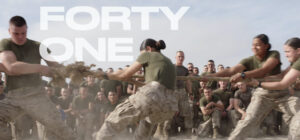 A group of military personnel in uniform are playing tug-of-war, pulling on a thick rope with determination. The background is filled with other service members watching and cheering. "FORTY ONE" is displayed prominently over the image, emphasizing their teamwork and spirit during this work in progress.