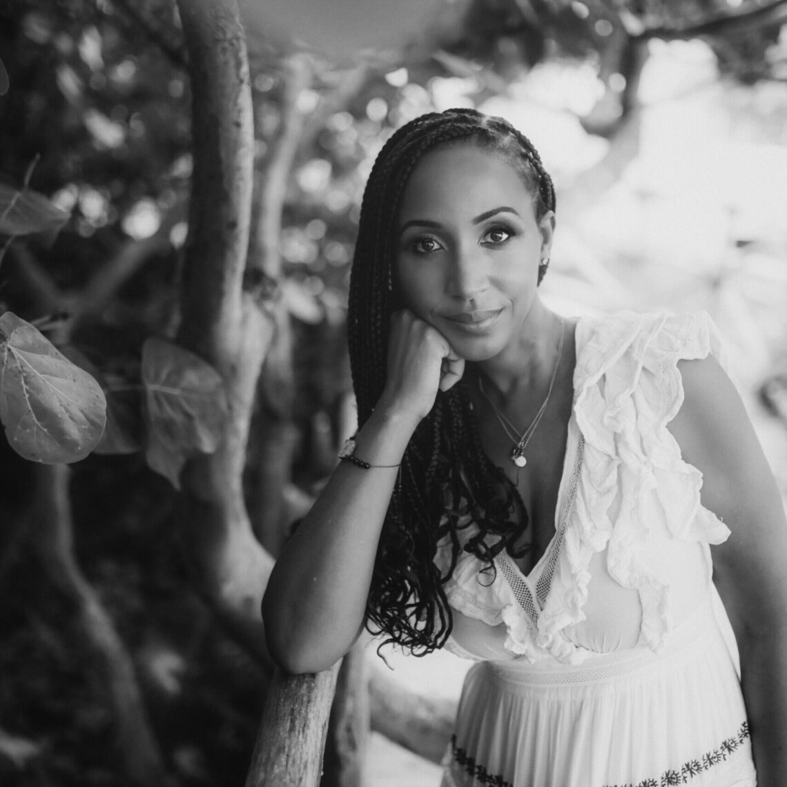 Black and white photo of a woman with long braided hair, leaning on a branch with her chin resting on her hand. She is wearing a sleeveless white dress adorned with ruffles. The background is blurred with foliage and tree branches, creating a serene, natural setting reminiscent of Berkeley Journalism's tranquil locales.