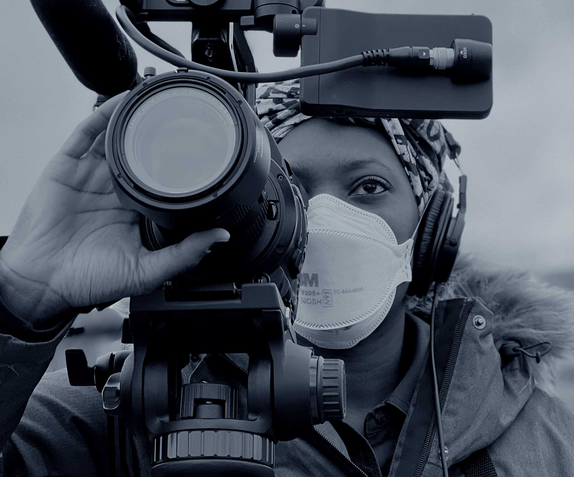 A person wearing a face mask, headphones, and a jacket operates a professional video camera with a large microphone attached. The person appears focused on the task, looking through the camera's viewfinder. The image is in black and white. Black woman journalist focusing a camera