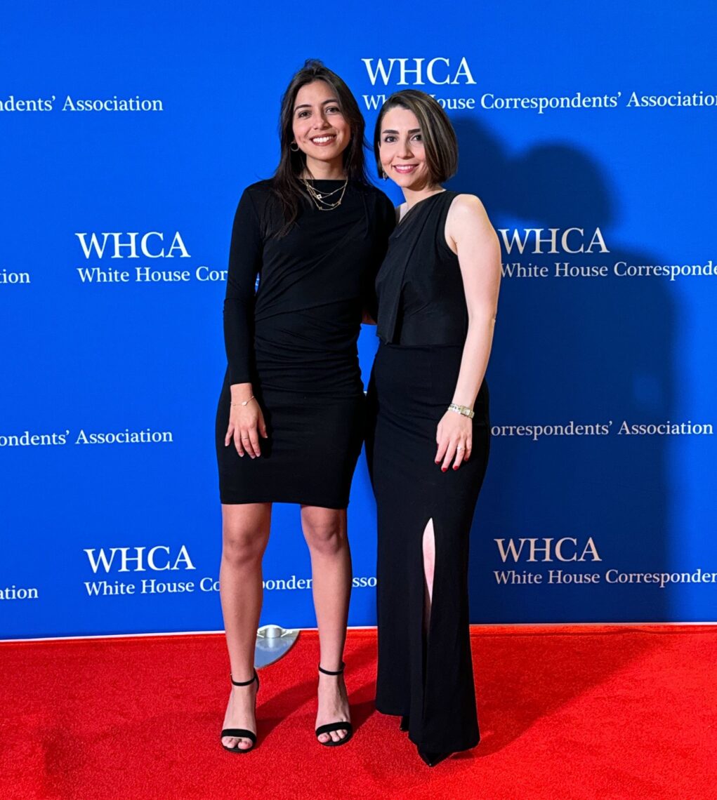 Photo of two women in black dresses standing close to one another on a red carpet in front of blue and white WHCA signage.
