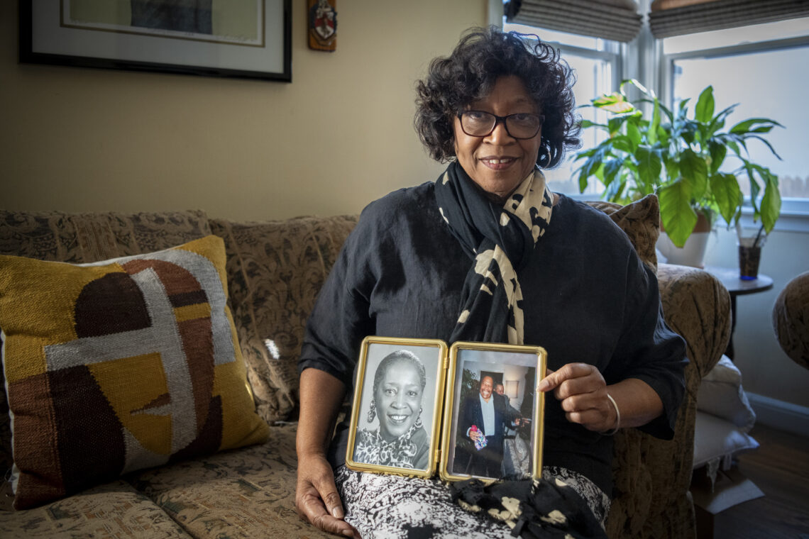 Gloria Brown, a Black woman with dark curly hair an thick-framed glasses sits on her couch holding a double-paned photo frame with a photo of her husband on the right side.