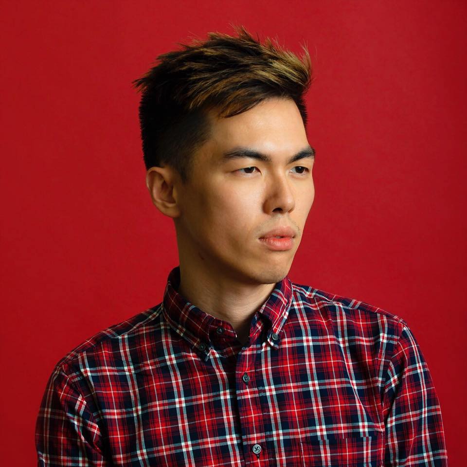 A person with short, styled hair is wearing a red and blue plaid shirt. They are looking off to the side against a solid red background.