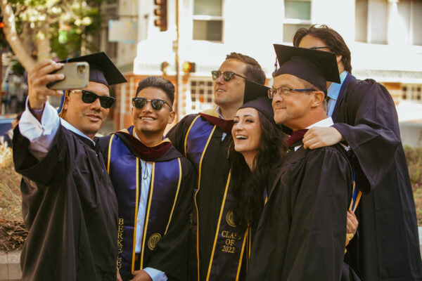 Group photo of 6 journalists wearing commencement gowns taking a selfie together smiling.
