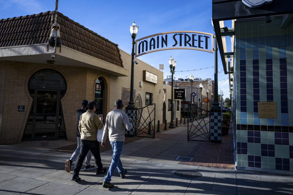 Photo of the arched Main Street sign between two streetlamps outside in downtown San Mateo. A group of three people are walking toward the signs. 