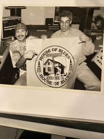Two men are holding up a large rectangular banner with the text "House of Blues Transmitter Network." The banner features an image of a house with musical notes and radio towers. The man on the left is wearing a bandana, and both are smiling.