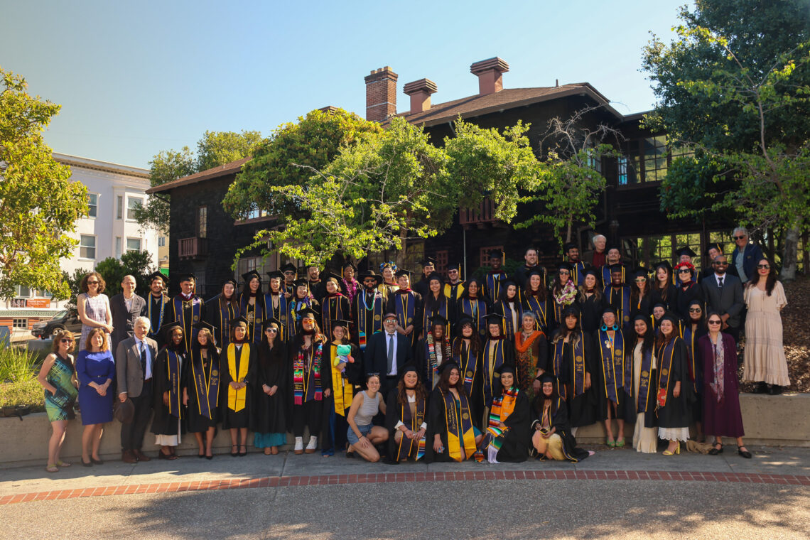 A large group of graduates in caps and gowns stand outside a building.
