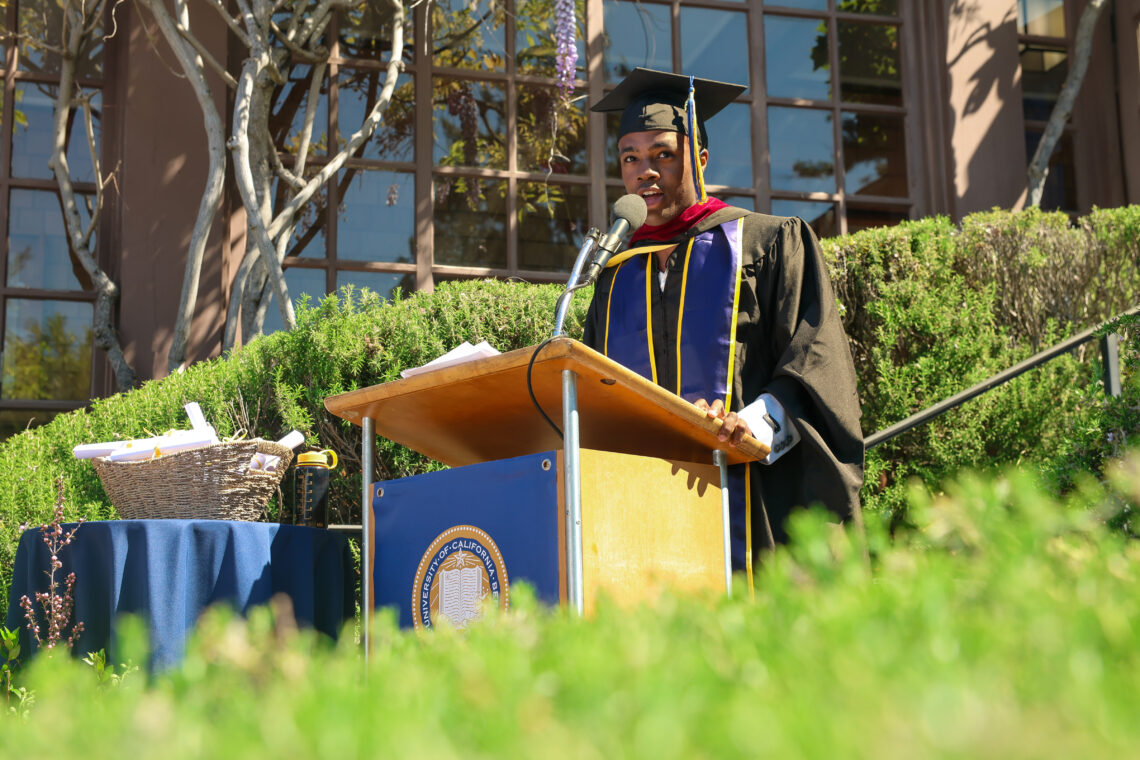 A graduate in a cap and gown speaks at a podium outdoors during the June 2024 commencement ceremony, holding the Dean's Letter with a proud smile.