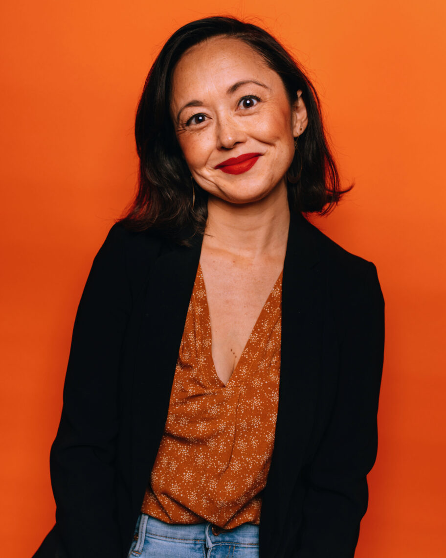 A woman smiles at the camera, wearing a black blazer and patterned blouse, standing against an orange background. 