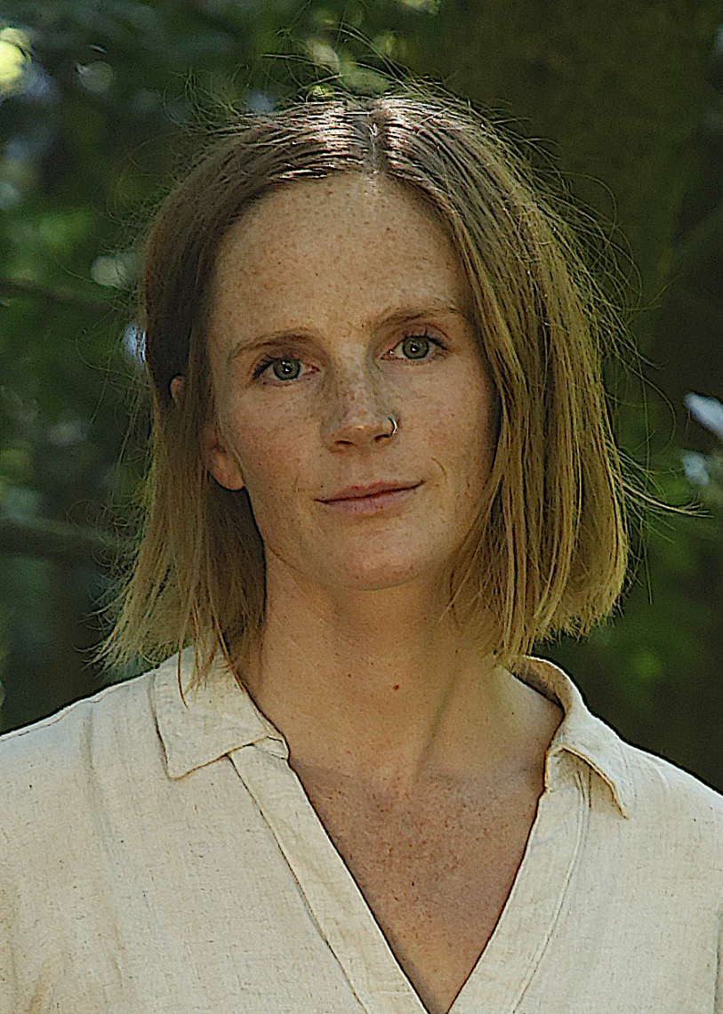 A woman with shoulder-length blonde hair and a nose ring stands outdoors, wearing a light-colored shirt. 