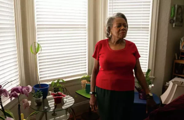 Vera Oliver, an older woman in a red shirt and black stands in front of the window in her San Francisco apartment. 