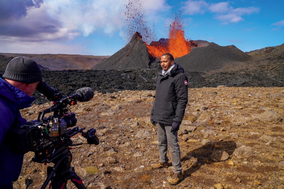 A man being filmed standing on rocky terrain with an erupting volcano in the background, capturing the raw power of nature. 