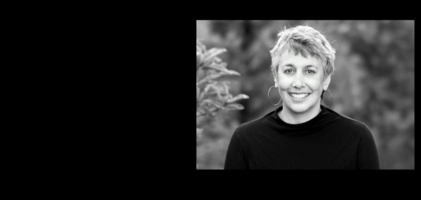 Black and white headshot of a smiling person with short hair and hoop earrings, wearing a dark sweater.