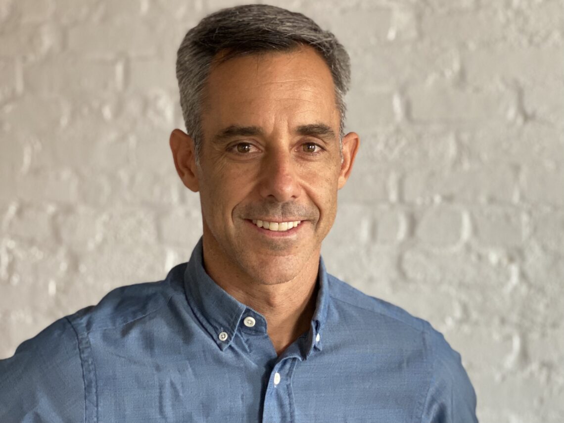 Man with short graying hair wears a blue button down shirt in front of a white brick background smiling.