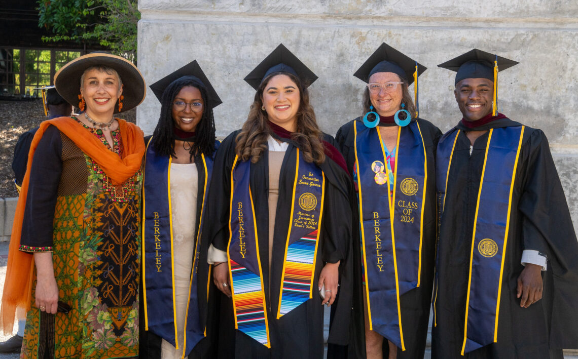 Five graduates in caps and gowns pose with Geeta Anand, a professor involved in teaching at Berkeley Journalism, who is dressed in colorful attire at the graduation ceremony.