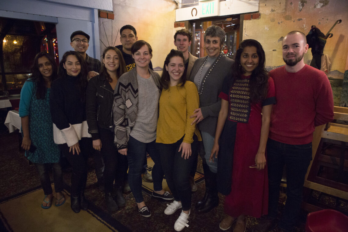 A group of eleven people, including Geeta Anand, the dean of Berkeley Journalism, standing and smiling in a dimly lit indoor setting.