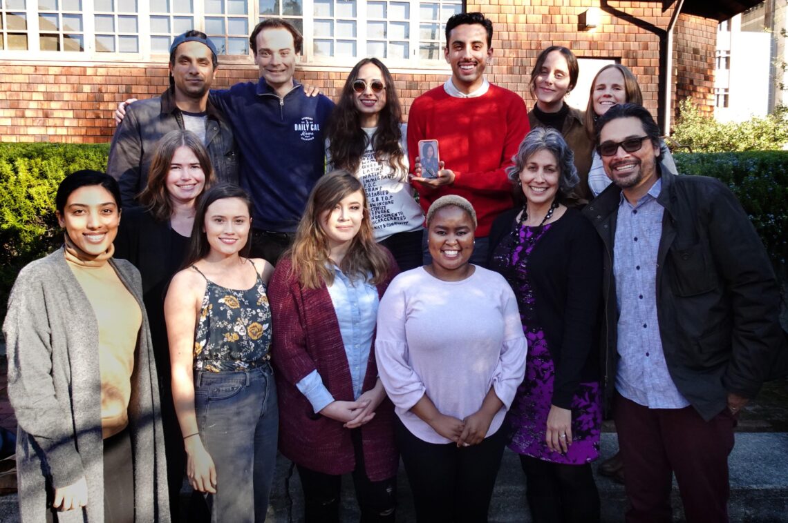 A group of thirteen people, including Geeta Anand, are posing together outside in front of a brick building. Some are smiling, others neutral. 