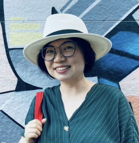 A person in a white hat and glasses smiles in front of a colorful wall mural.