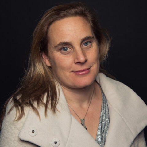 Jessica Garrison, a woman with light brown hair wearing a white coat and a necklace, poses gracefully in front of a dark background.