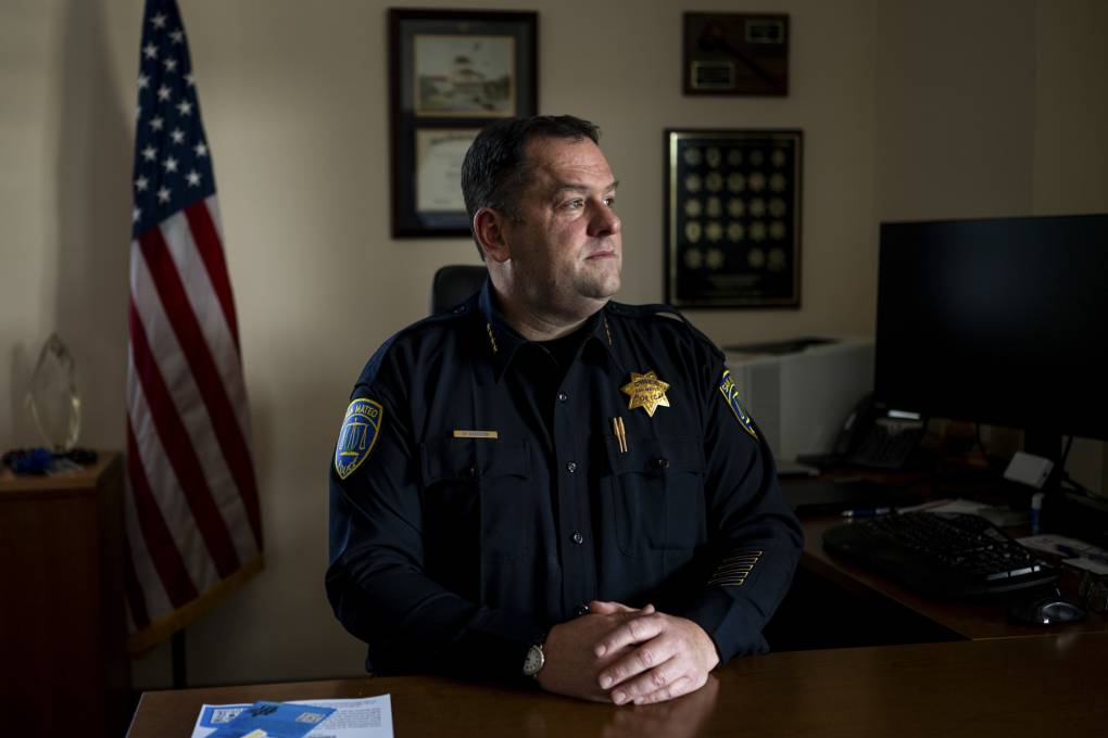 A police officer in uniform stands with hands folded in an office, exuding compassion. An American flag and computer are visible in the background.