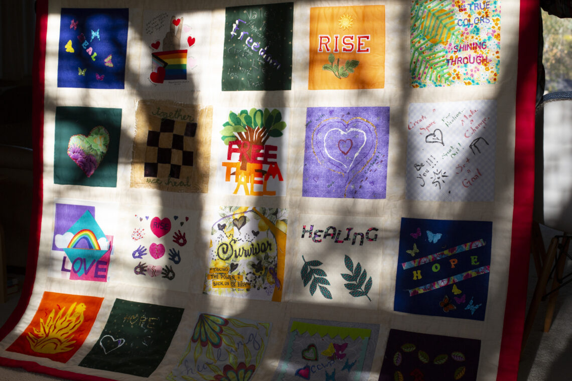 A colorful patchwork quilt with various square panels showing designs and words like "Rise," "Freedom," "Healing," and honoring Forced Sterilization Survivors.