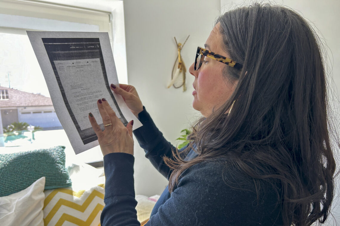 A woman with long dark hair and glasses reads a document while standing near a bright window, reflecting on the stories of forced sterilization survivors.