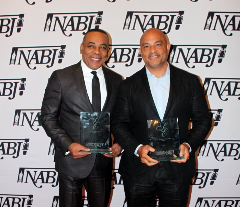 Two men in suits hold awards in front of a step-and-repeat backdrop adorned with the NABJ logo, celebrating their achievements at the National Association of Black Journalists' Salute to Excellence Awards.