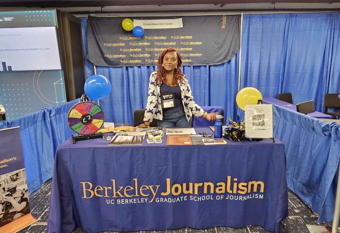 A woman stands behind a Berkeley Journalism booth with a colorful spin wheel and informational materials.