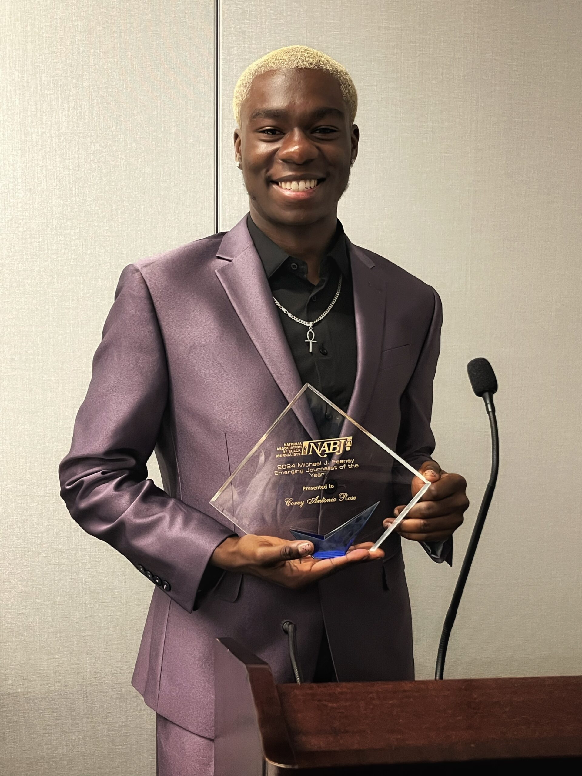 A person in a purple suit smiles while holding a glass award near a podium with a microphone.