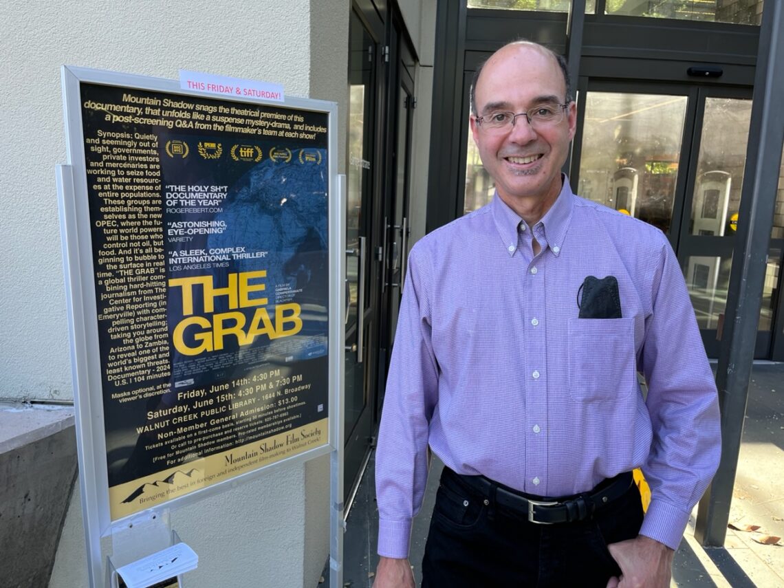 Man wearing glasses and a button down shirt stands next to a theater poster of The Grab smiling.