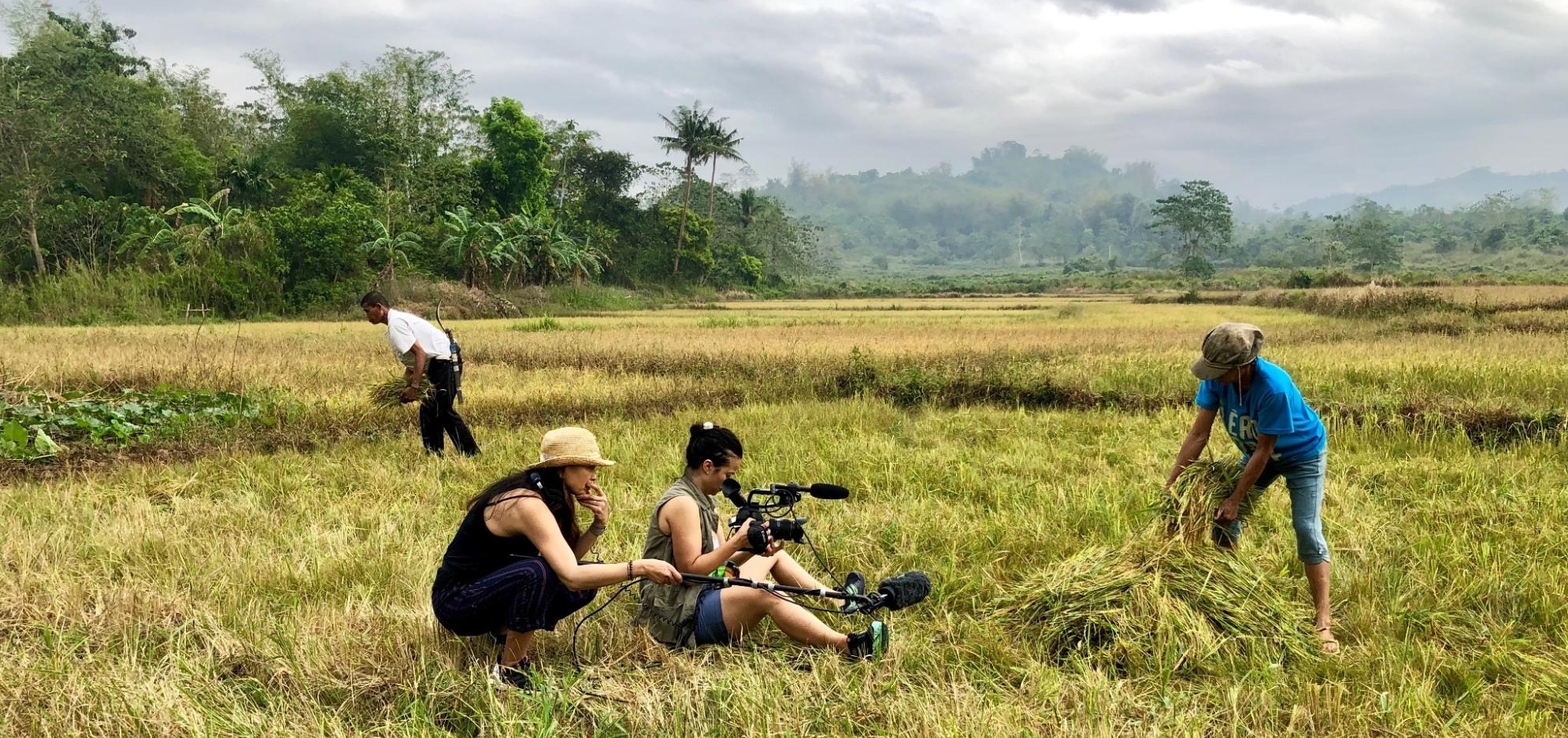 People filming in a field with cameras for a climate journalism program while others work on the crops, green hills and cloudy sky in the background.