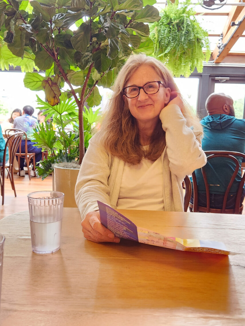 A woman with long medium brown wavy hair wearing glasses sits at a table in an outdoor restaurant smiling and wearing a cream colored sweater.