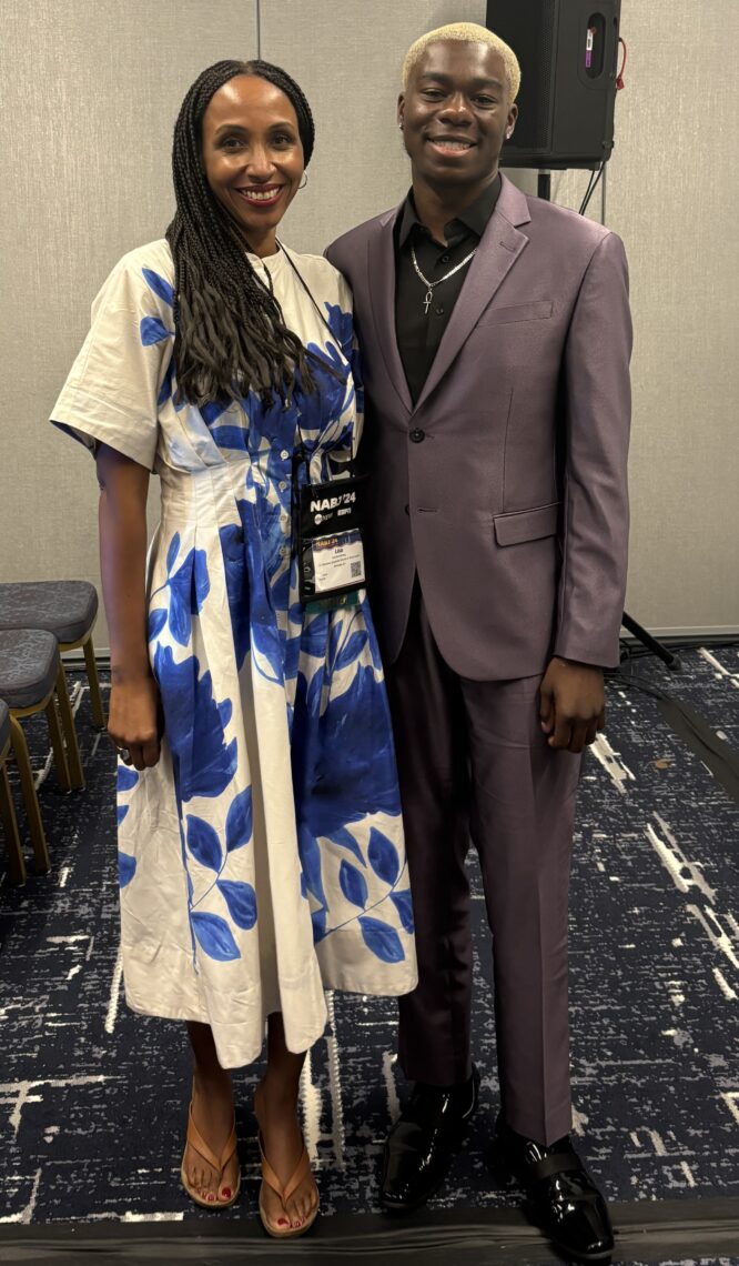 Two people smiling; a woman in a white dress with blue floral patterns, and a man in a purple suit.