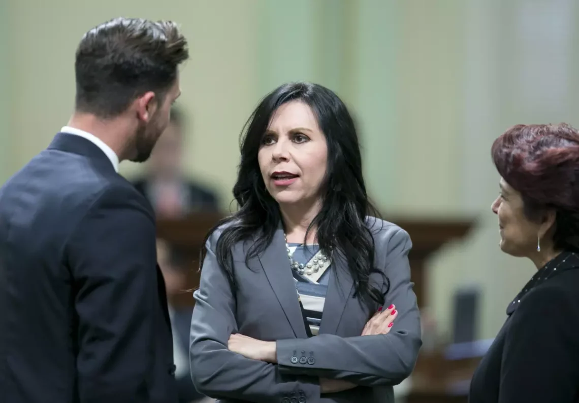 Blanca Rubio, a petite woman with long straight black hair, stands with her arms crossed speaking with two other people in suits. 